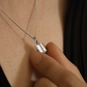Peut inclure: Un collier argenté doté d'un petit pendentif en pierre translucide rectangulaire maintenu par une monture métallique simple. Le pendentif est suspendu à une fine chaîne en argent à maillons délicats, mettant en valeur une pierre facettée claire.