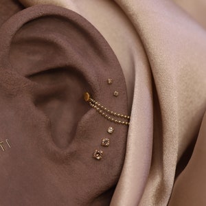 May include: A close-up of a person's ear with multiple small diamond earrings and a gold chain earring. The earrings are set against a brown background.