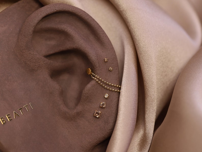May include: A close-up of a person's ear with multiple small diamond earrings and a gold chain earring. The earrings are set against a brown background.
