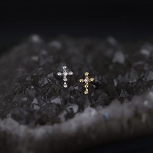 May include: Two small cross-shaped earrings. One is silver-colored, the other is gold-colored. Each earring has a small, clear gemstone at the center of the cross. The earrings are displayed on a dark, textured surface.