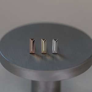 May include: Three geometric stud earrings in rose gold, gold, and silver tones. The earrings are rectangular with a triangular top. The word "BEATTI" is printed in white below the earrings. The earrings are displayed on a silver surface.
