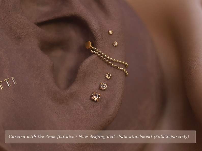 May include: Gold ear jewelry with a small flat disc and a chain with small gold balls hanging from it. There are also several small clear gemstones on the ear.