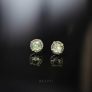 May include: A pair of stud earrings with light green gemstones set in a gold-colored metal. The earrings are round with a textured bezel setting. The image is set against a dark background with the word "BEATTI" at the bottom.