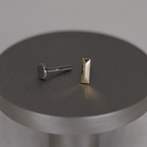 May include: Two pieces of jewelry on a gray surface. One piece is a silver-colored labret stud with a flat, round disc. The other is a gold-colored, rectangular stud. The word "BEATTI" is printed on the surface.