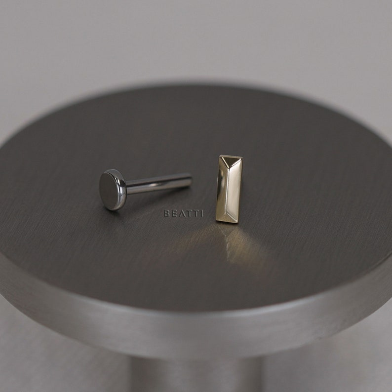 May include: Two pieces of jewelry on a gray surface. One piece is a silver-colored labret stud with a flat, round disc. The other is a gold-colored, rectangular stud. The word "BEATTI" is printed on the surface.