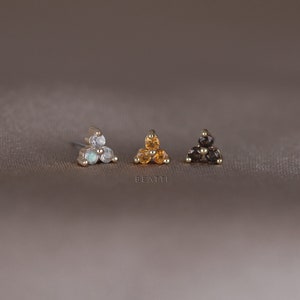 May include: Three small stud earrings with a triangular design. One earring features opalescent stones, another has yellow stones, and the third has dark grey stones. The earrings are set on a gold-coloured metal. The word "BEATTI" is visible.