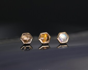 May include: Three gold hexagonal earrings with different colored gemstones. The earrings are on a dark gray surface. The gemstones are a light brown, a dark brown, and a clear iridescent stone.