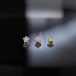 May include: Three flower-shaped stud earrings in silver, rose gold, and gold. Each earring features a central gemstone. The earrings are displayed on a reflective surface, creating mirrored reflections of the jewelry.