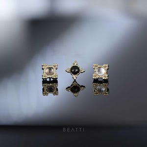 May include: Three gold-coloured, square-shaped jewellery pieces with ornate edges and a central gemstone. One has a black gem, and the others have clear gems. The pieces are displayed on a reflective surface.