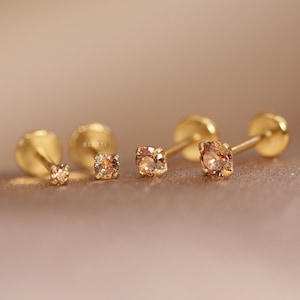 May include: Four gold stud earrings with small brown gemstones. The earrings are arranged in a row, with the largest earring on the right and the smallest earring on the left.