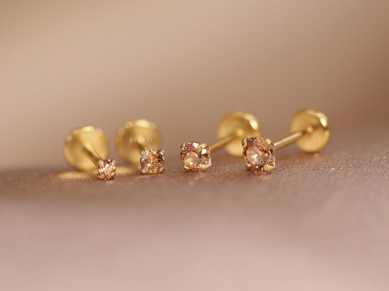 May include: Four gold stud earrings with small brown gemstones. The earrings are arranged in a row, with the largest earring on the right and the smallest earring on the left.