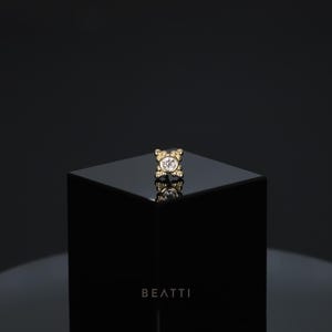May include: A gold-colored, teddy bear-shaped stud earring with a clear, round gemstone in the center. The earring is displayed on a black, glossy surface. The word "BEATTI" is printed in white at the bottom.