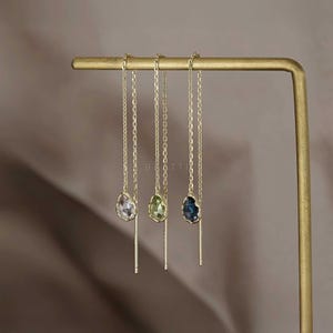 May include: Three gold-toned chain earrings with teardrop-shaped gemstones in clear, green, and blue hues. Each earring features a delicate chain and a straight bar. The earrings are displayed on a gold-colored stand.