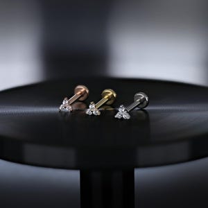 May include: Three flat-back earrings with a triangular cluster of small, clear gemstones. The earrings are in rose gold, yellow gold, and silver tones. The earrings are displayed on a black surface.