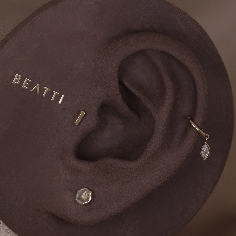 May include: Close-up of an ear adorned with gold earrings. The ear has multiple piercings, including a small hoop with a dangling teardrop-shaped gem, a bar stud, and a hexagonal stud. The word "BEATTI" is embossed on the surface.