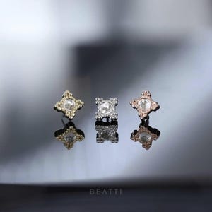 May include: Three stud earrings with a four-petal design and a central clear stone. The earrings are in gold, silver, and rose gold tones, each reflecting light. The brand name "BEATTI" is at the bottom.