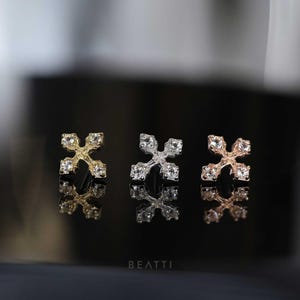 May include: Three stud earrings in gold, silver, and rose gold, each with a cross design and clear gemstones. The earrings are displayed on a reflective black surface, with the brand name "BEATTI" visible.
