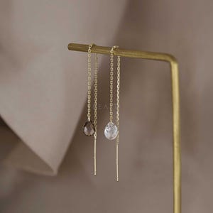 May include: A pair of gold-coloured threader earrings. One earring has a smoky quartz teardrop, the other a clear crystal teardrop. Both hang from delicate chains and are displayed on a gold-coloured earring stand.
