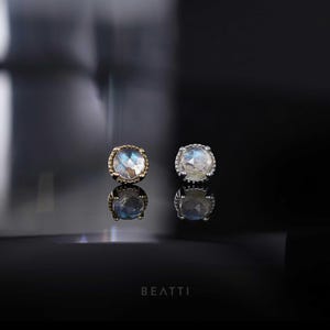 May include: Two round stud earrings with iridescent, light blue and white stones. One earring has a gold setting, and the other has a silver setting. The earrings are displayed on a reflective black surface. The word "BEATTI" is at the bottom.