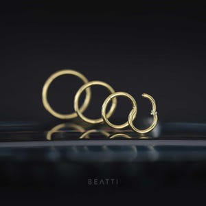 May include: Four gold-colored hoop earrings of varying sizes are arranged in a row, with one open. The earrings are displayed against a dark background with a reflective surface. The word "BEATTI" is at the bottom.