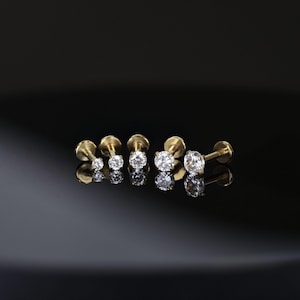May include: A selection of gold labret studs, each featuring a clear, round gemstone. The studs are displayed in a line, with varying gemstone sizes, reflecting light on a dark, glossy surface. These are designed for lip or ear piercings.