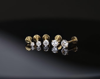 May include: A collection of gold-colored labret studs with clear, round-cut gemstones. The studs are arranged in a row, each with a different size gemstone, reflecting light against a dark, glossy surface. The jewelry is designed for lip or ear piercings.