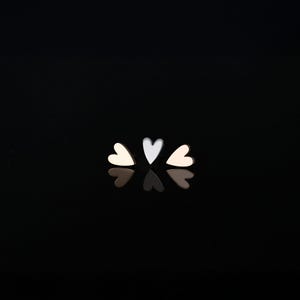 May include: Three heart-shaped stud earrings in a row, with a reflective surface below. The earrings are in three colors: cream, silver, and pale gold. The background is solid black.