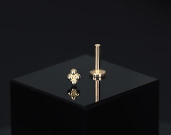 May include: Two gold-colored body jewelry pieces. One piece features a cluster of four small spheres, and the other has a flat base with a post. Both pieces are displayed on a black surface against a dark background.