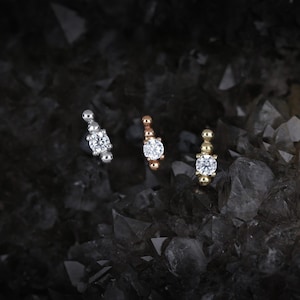 May include: Three stud earrings in silver, rose gold, and gold. Each earring features a central, round, clear gemstone, surrounded by smaller, spherical accents. The earrings are displayed against a dark, textured background, creating a contrast that highlights their sparkle.
