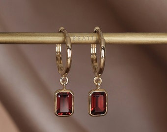 May include: Gold hoop earrings with a dangling red rectangular gemstone. The earrings are on a gold stand with the word "BEATTIE" printed on it.