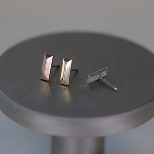 May include: Three pairs of stud earrings in different metallic finishes: rose gold, gold, and silver. Each earring has a rectangular bar shape with a faceted design. The earrings are displayed on a gray, circular surface.