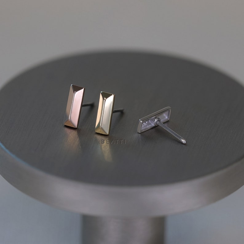 May include: Three pairs of stud earrings in different metallic finishes: rose gold, gold, and silver. Each earring has a rectangular bar shape with a faceted design. The earrings are displayed on a gray, circular surface.