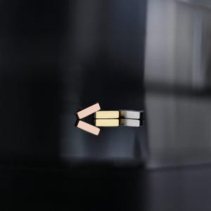 Peut inclure: Une collection de boucles d'oreilles rectangulaires en or rose, or et argent. Les boucles d'oreilles sont disposées sur une surface noire réfléchissante, avec leurs reflets en miroir en dessous. Le design minimaliste suggère une esthétique de bijoux moderne.