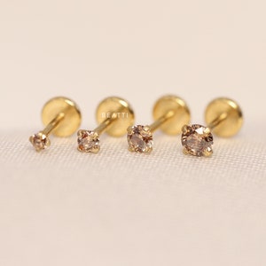 May include: Four gold stud earrings with small, clear, round gemstones. The earrings are arranged in a row on a white surface.