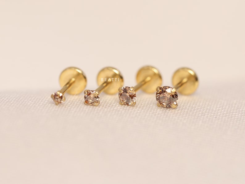 May include: Four gold stud earrings with small, clear, round gemstones. The earrings are arranged in a row on a white surface.