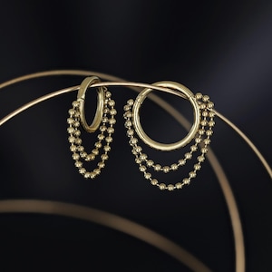 May include: A pair of gold hoop earrings with dangling beaded chains. The earrings are presented against a dark backdrop, accentuating their polished finish and intricate design. The beaded chains add a touch of elegance.