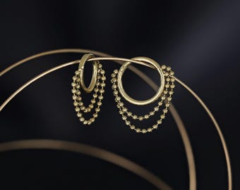May include: A pair of gold hoop earrings with dangling beaded chains. The earrings are displayed against a dark background, highlighting their polished finish and intricate design. The beaded chains add a touch of elegance.