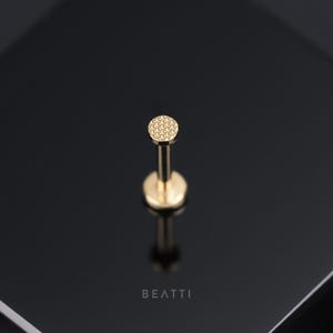 May include: Gold body jewelry with a round, textured top. The jewelry is on a black background.