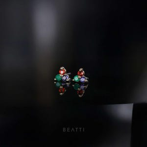 May include: A pair of stud earrings with a cluster of colorful gemstones. Each earring features red, green, and purple stones, set in a gold-toned metal. The earrings are displayed on a reflective black surface.