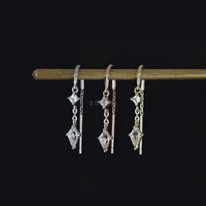 May include: Three pairs of dangling earrings with a geometric design. Each earring features a small square gem, a chain or bar, and a larger, diamond-shaped gem. The earrings are silver, rose gold, and gold, displayed against a black background.