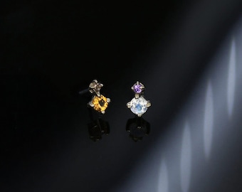 May include: A pair of stud earrings with a silver setting. One earring features a yellow square-cut gemstone, while the other has a white round gemstone topped with a small purple gemstone. The earrings are displayed against a dark background.