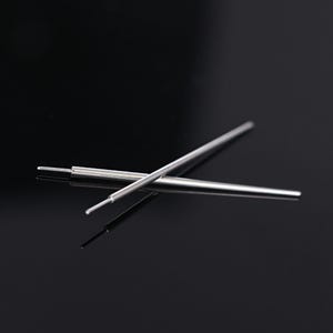 May include: Two silver metal pen refills are crossed against a black background. The refills are cylindrical with a fine tip. The image is a close-up shot, highlighting the metallic sheen and the precision of the pen refills.