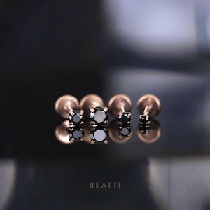 May include: Four rose gold stud earrings with black gemstones are shown on a reflective surface. The earrings vary in size, with the largest in the center. The word "BEATTI" is printed at the bottom.