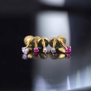 May include: Four gold-coloured stud earrings, each featuring a small gemstone in shades of pink, purple, and clear. The earrings are displayed on a reflective surface. The word 'BEATTI' is at the bottom.