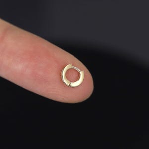 Könnte beinhalten: Ein kleiner, goldfarbener Creolen-Ohrring. Der Ohrring hat ein einfaches, kreisförmiges Design mit einem Klappverschluss. Der Ohrring wird auf einer Fingerspitze vor dunklem Hintergrund gezeigt.