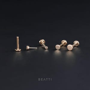 May include: Four gold-toned body piercing studs with round ends. The studs are arranged in a row on a black surface. The word "BEATTII" is printed in white on the surface.