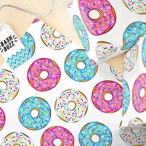 K&ouml;nnte beinhalten: Wei&szlig;es Geschenkpapier mit einem sich wiederholenden Muster aus bunten Donuts mit Streuseln. Die Donuts sind rosa, blau und wei&szlig; mit bunten Streuseln.