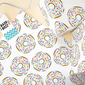 K&ouml;nnte beinhalten: Wei&szlig;es Geschenkpapier mit einem sich wiederholenden Muster aus bunten Donuts mit Streuseln. Die Donuts sind hellblau mit wei&szlig;em Zuckerguss und bunten Streuseln. Das Papier hat den Text "Bash Buzz" auf einem blau-wei&szlig;en Etikett.