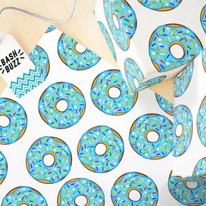 K&ouml;nnte beinhalten: Wei&szlig;es Geschenkpapier mit einem sich wiederholenden Muster aus blauen Donuts mit bunten Streuseln. Die Donuts haben eine wei&szlig;e Mitte und einen braunen Umriss.