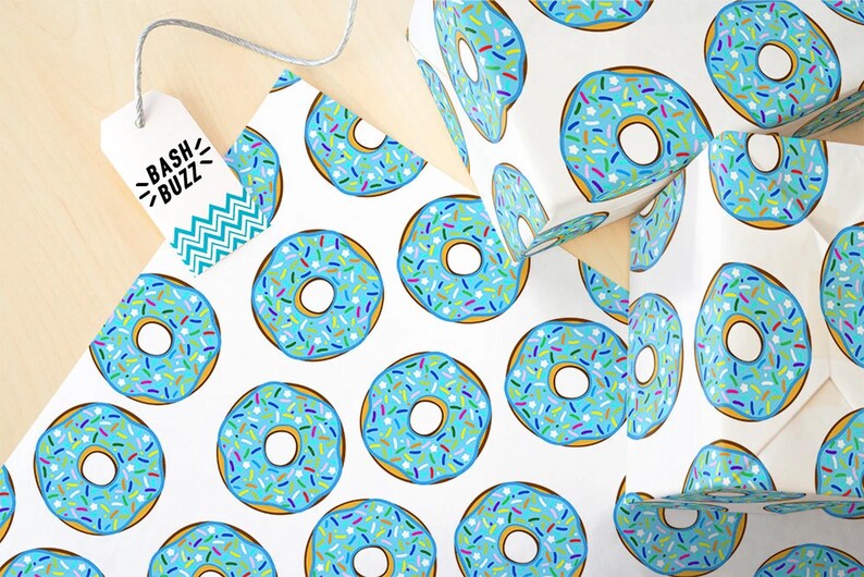 K&ouml;nnte beinhalten: Wei&szlig;es Geschenkpapier mit einem sich wiederholenden Muster aus blauen Donuts mit bunten Streuseln. Die Donuts haben eine wei&szlig;e Mitte und einen braunen Umriss.